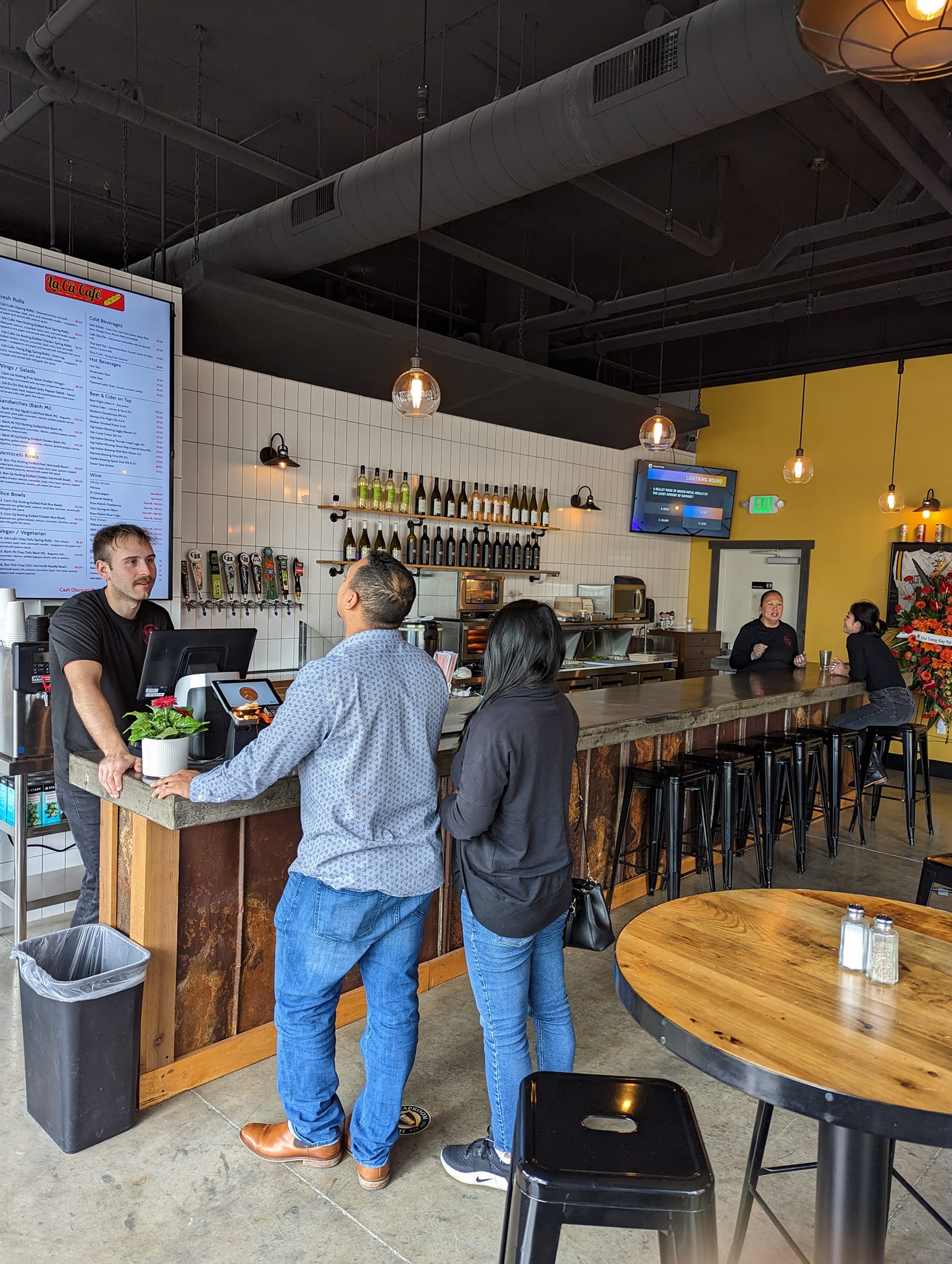 Interior of La Ca Cafe in Tacoma showing customers ordering at counter with visible menu board, industrial-casual decor with pendant lighting, beer taps, and wooden bar seating - Vietnamese restaurant near PLU campus