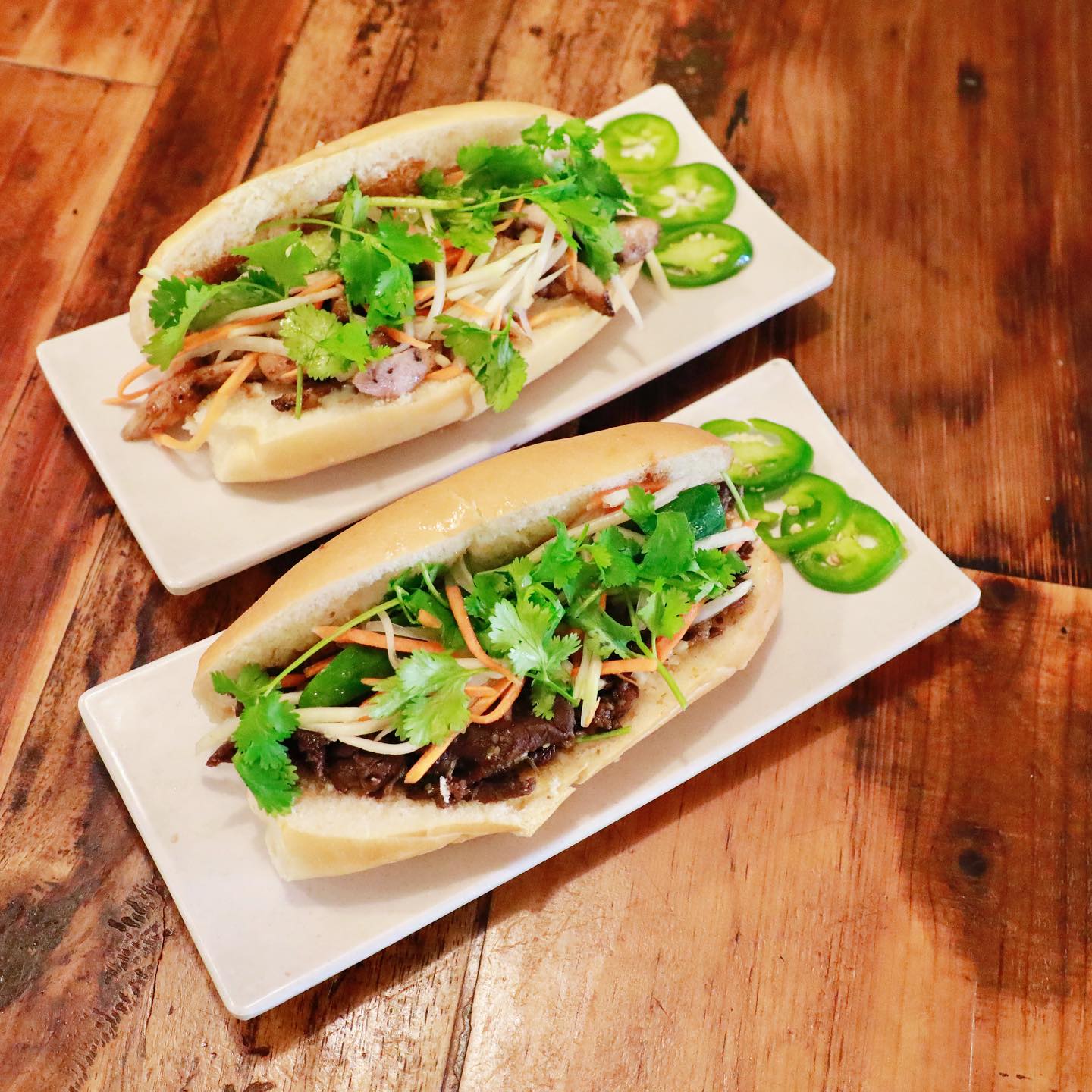 Two Vietnamese bánh mì sandwiches at La Cà Café Tacoma featuring grilled pork and grilled chicken on fresh baguettes with pickled vegetables, fresh cilantro, jalapeños, and traditional garnishes on white plates with rustic wooden table